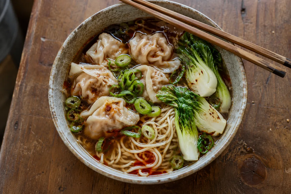 Top-down view of wonton noodle soup in a ceramic bowl with dark beef broth, red chili oil, pleated pork wontons, bok choy, noodles and scallions with chopsticks