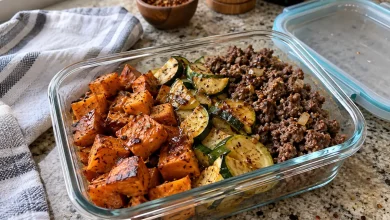 A beef sweet potato bowl in a clear glass meal-prep container with roasted sweet potato cubes, zucchini slices, browned ground beef and a honey drizzle