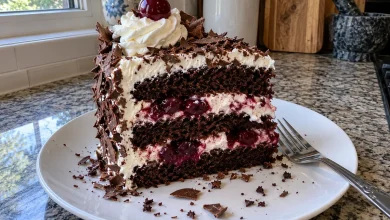 Slice of black forest cake on a white plate showing chocolate sponge layers, whipped cream and sour cherries with chocolate shavings on top